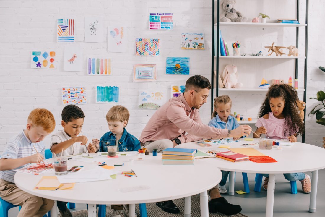 teacher and interracial children drawing pictures together at tables in classroom